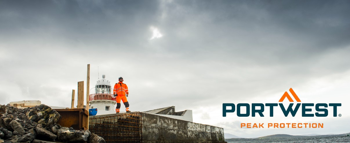 Een arbeider, gekleed in oranje hoge zichtbaarheidskleding, staat op een rotsachtige bouwplaats naast een vuurtoren, met het Portwest-logo en de slogan 'Peak Protection' op de foto.