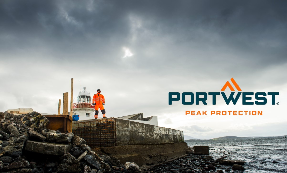 Een arbeider, gekleed in oranje hoge zichtbaarheidskleding, staat op een rotsachtige bouwplaats naast een vuurtoren, met het Portwest-logo en de slogan 'Peak Protection' op de foto. Er is een link die leidt naar onze multinorm werkkleding.