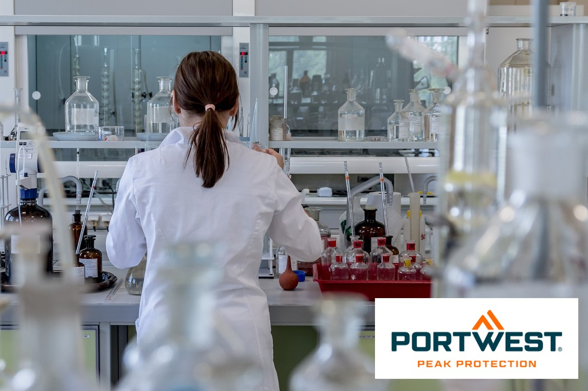 Een vrouw met donkerbruin achterover gebonden haar draagt ​​een witte laboratoriumjas en werkt in een modern laboratorium met diverse laboratoriumapparatuur en chemicaliënflessen. Rechtsonder in de afbeelding staat het logo “Portwest Peak Protection”. Er is een link die naar al onze laboratoriumjassen leidt.