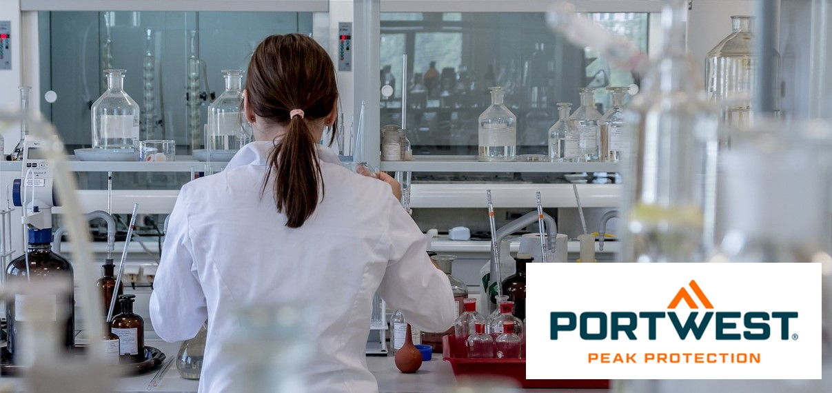 Een vrouw met donkerbruin achterover gebonden haar draagt ​​een witte laboratoriumjas en werkt in een modern laboratorium met diverse laboratoriumapparatuur en chemicaliënflessen. Rechtsonder in de afbeelding staat het logo “Portwest Peak Protection”.