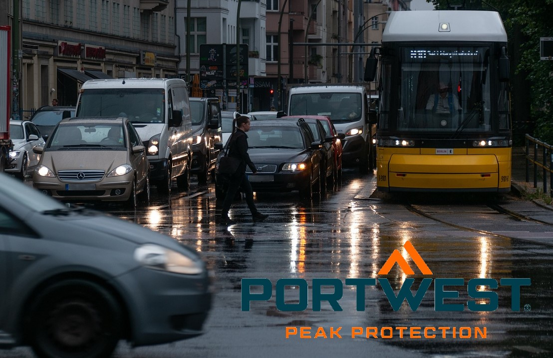 Straatbeeld in de regen. Op een kruispunt staan ​​talloze auto's en bussen met ingeschakelde koplampen geparkeerd. Het bedrijfslogo van Portwest in oranje en blauw bevindt zich in de rechter benedenhoek van de foto. Er is een link die naar onze regenjassen leidt.