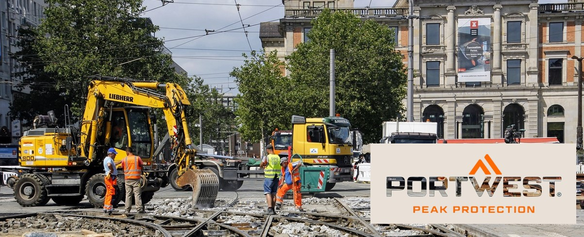 Bouwvakkers in oranje veiligheidskleding werken op een bouwplaats in de stad. Op de achtergrond zijn graafmachines, vrachtwagens en een historisch gebouw te zien. Onderaan de afbeelding zijn het Portwest-logo en de slogan "Peak Protection" weergegeven.