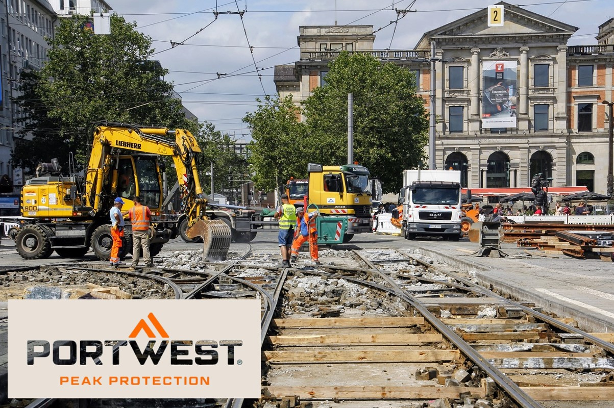 Bouwvakkers in oranje veiligheidskleding werken op een bouwplaats in de stad. Op de achtergrond zijn graafmachines, vrachtwagens en een historisch gebouw te zien. Onderaan de afbeelding zijn het Portwest-logo en de slogan "Peak Protection" weergegeven. Er is een link die naar de artikelpagina van de koelkap leidt.